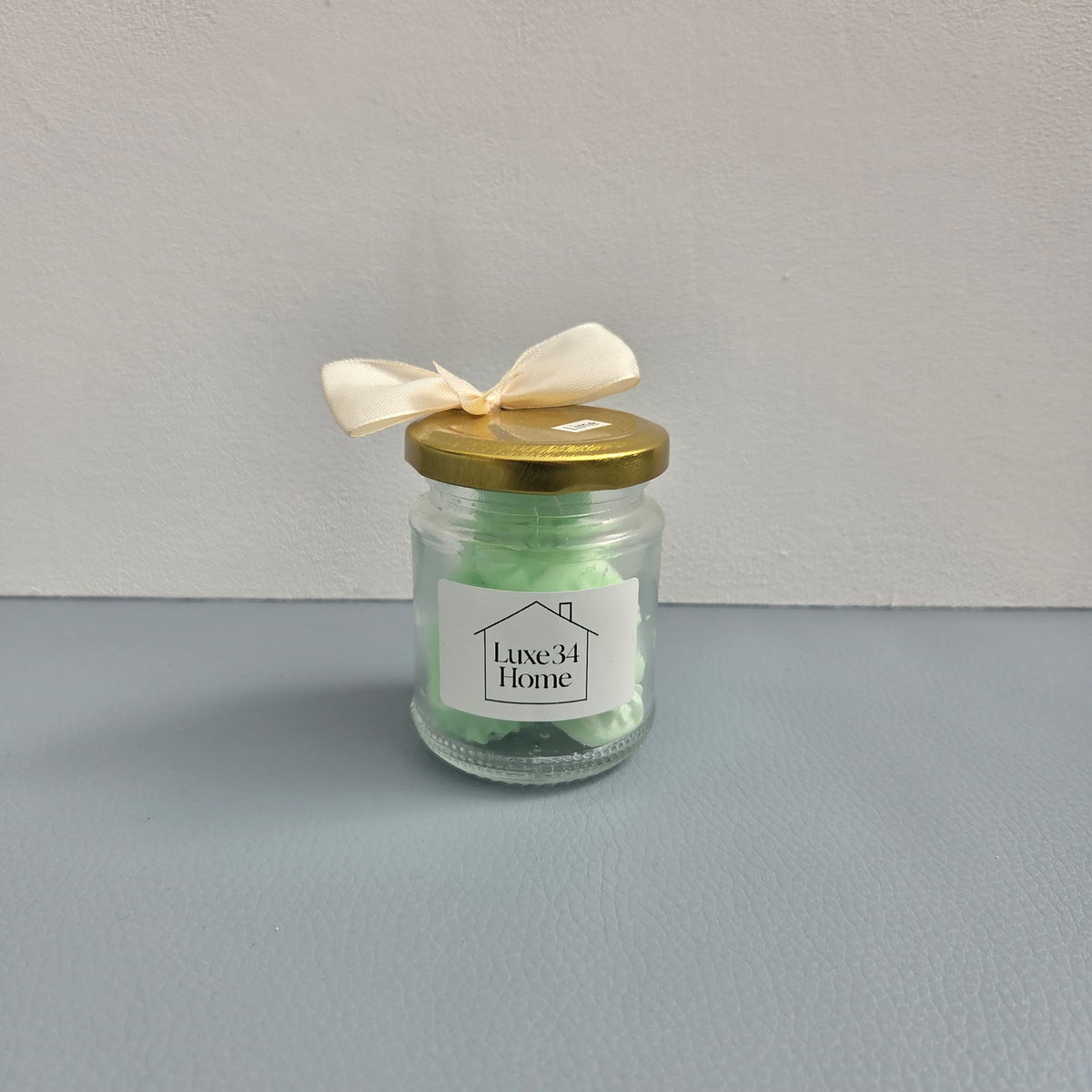 A small clear glass jar candle with a gold metal lid, tied with a cream satin ribbon. The candle wax inside is light green, and the front features a white label with the brand name 'Luxe34 Home'.