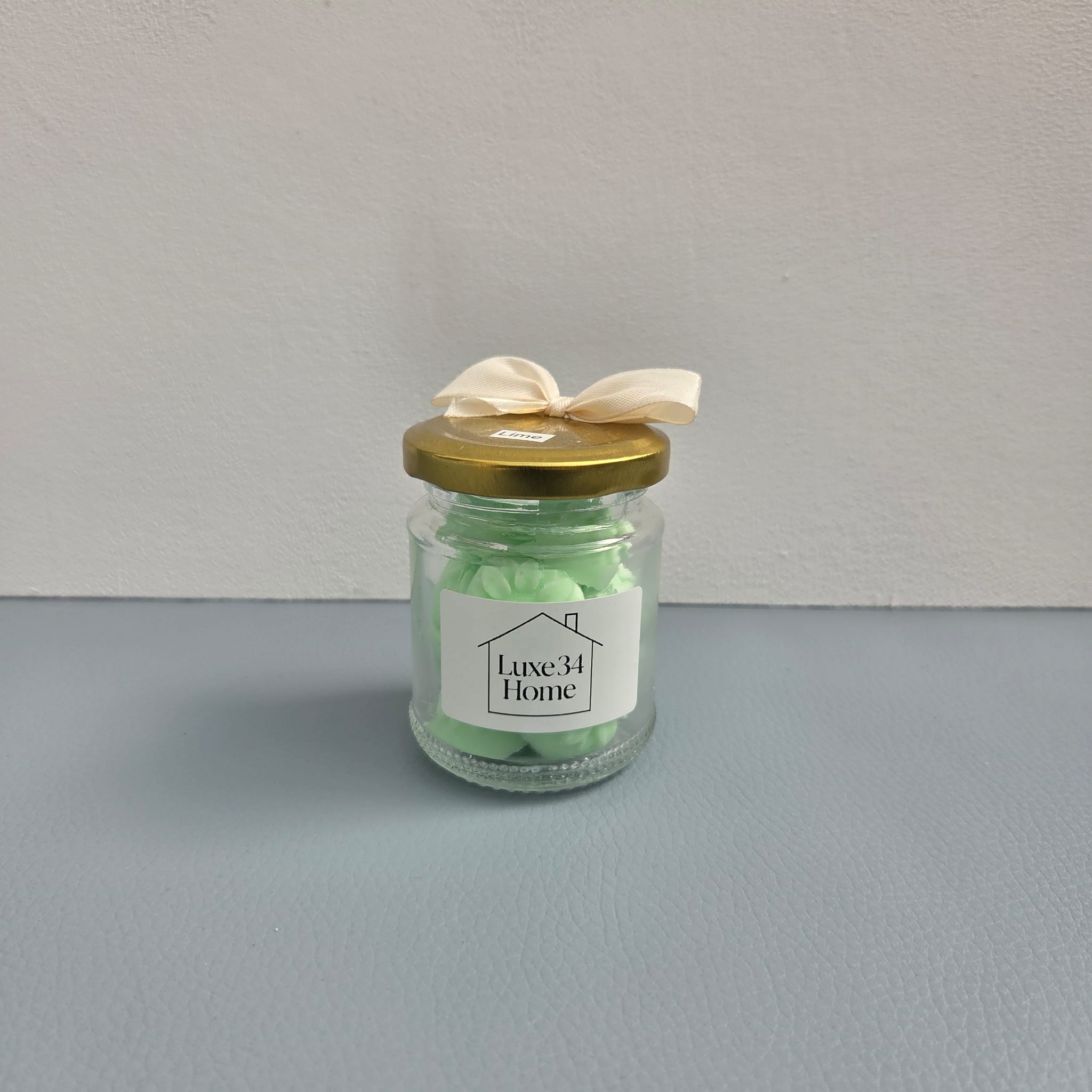A small clear glass jar candle with pale green wax, a gold metal lid, and a cream ribbon bow on top. The front label reads 'Luxe 34 Home'.