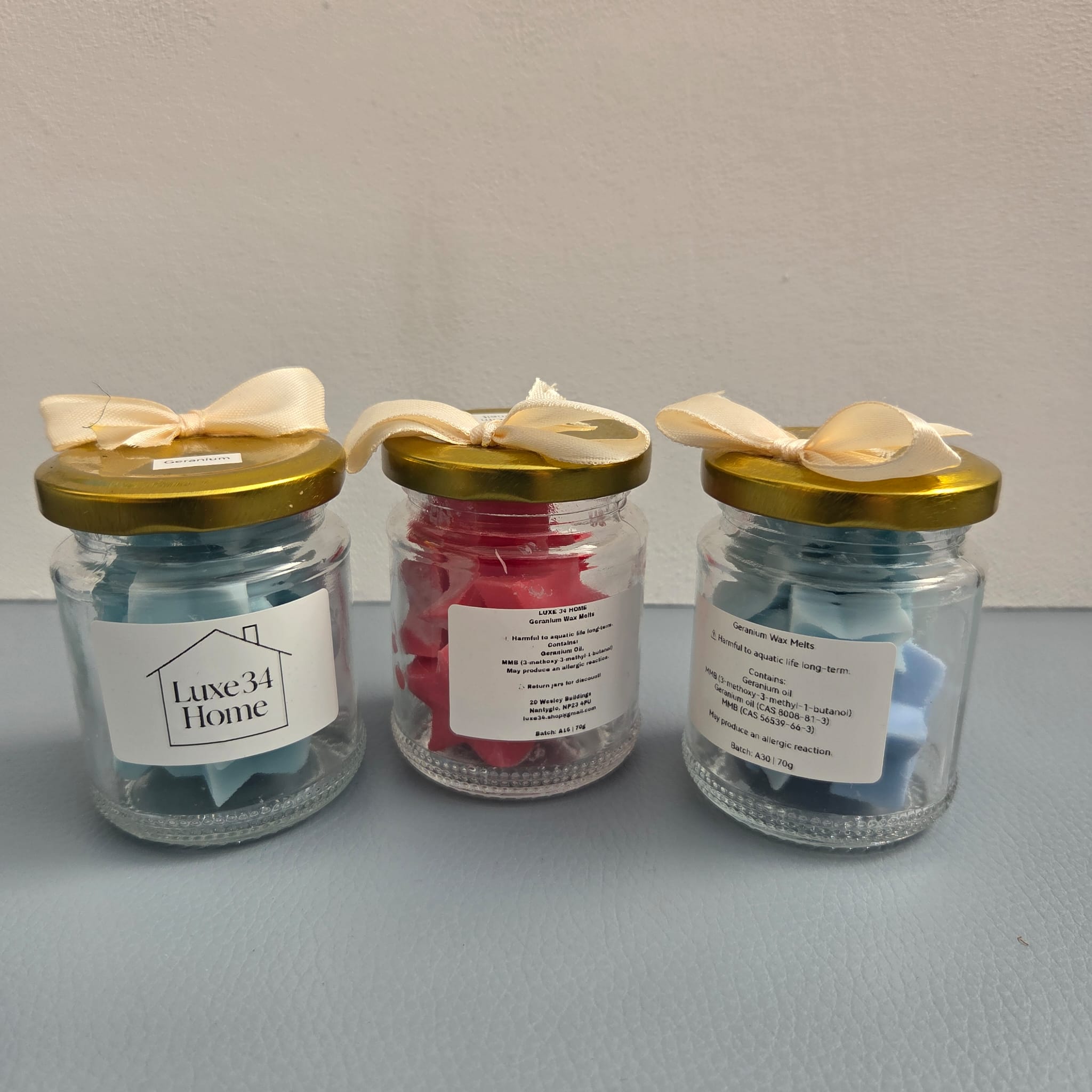 Three glass jars with gold lids and cream ribbon bows, containing wax melts, arranged on a light blue surface against a white wall. The left jar is labeled 'Luxe34 Home', and the middle and right jars have product labels but the text is not fully legible.