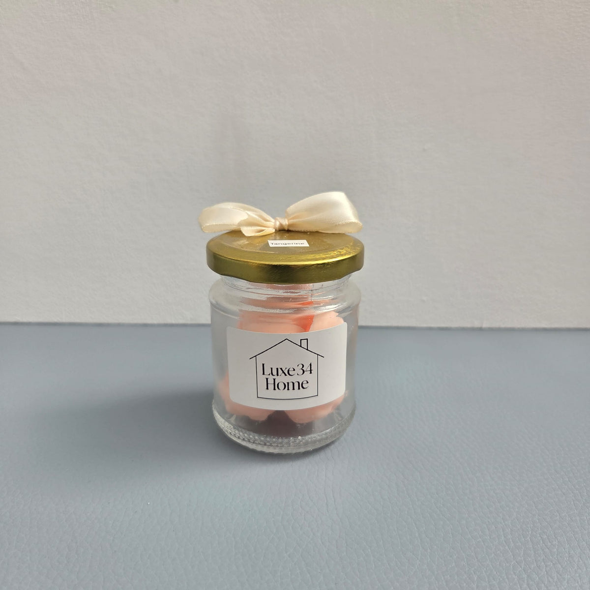 A small clear glass jar candle with a gold lid and a cream ribbon bow, featuring a white label that reads 'Luxe34 Home'. The candle wax inside is a soft peach-pink color, and the bottom of the jar contains dark brown wicks.