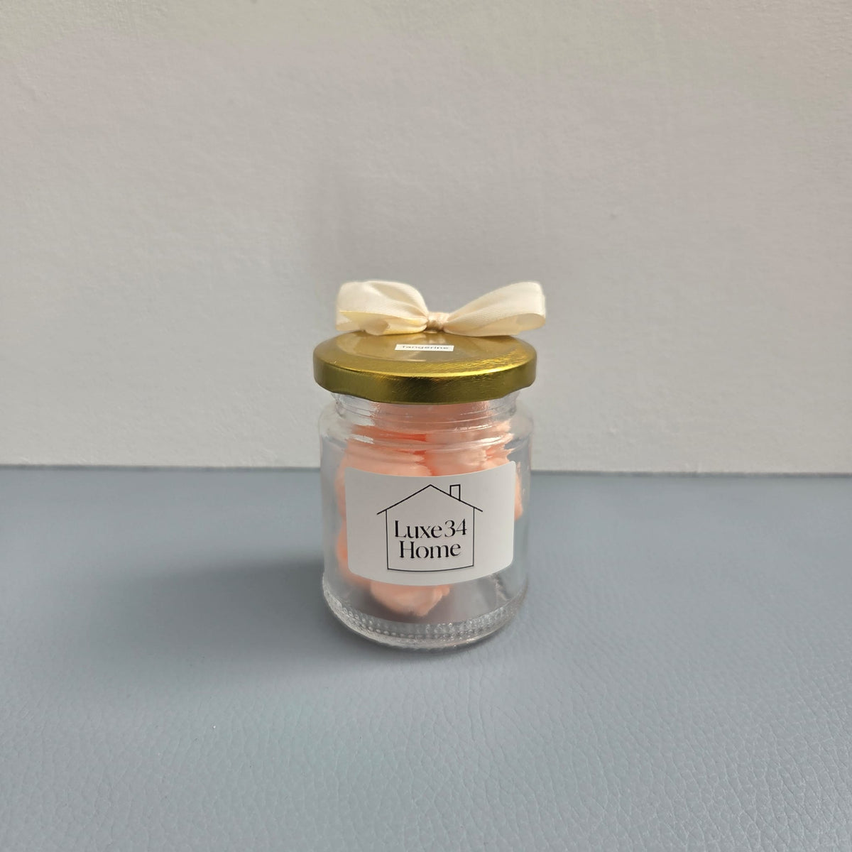 A clear glass jar candle with pale peach wax, a gold metal lid, and a cream satin bow, featuring a white label that reads 'Luxe34 Home'.