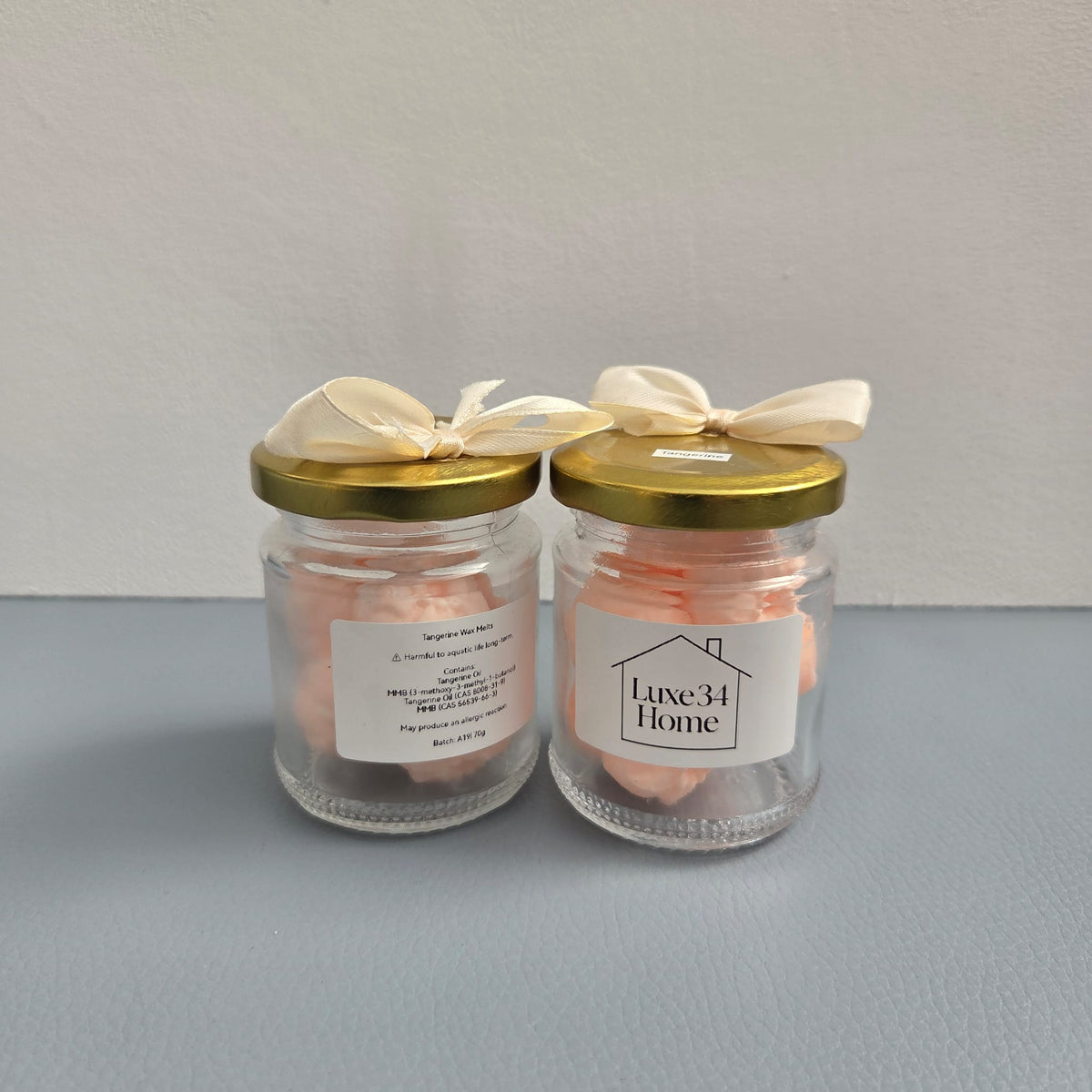Two clear glass jars with gold lids, filled with pale pink bath salts, tied with cream ribbons, and displayed on a gray surface against a light gray background. One label reads 'Luxe34 Home'.