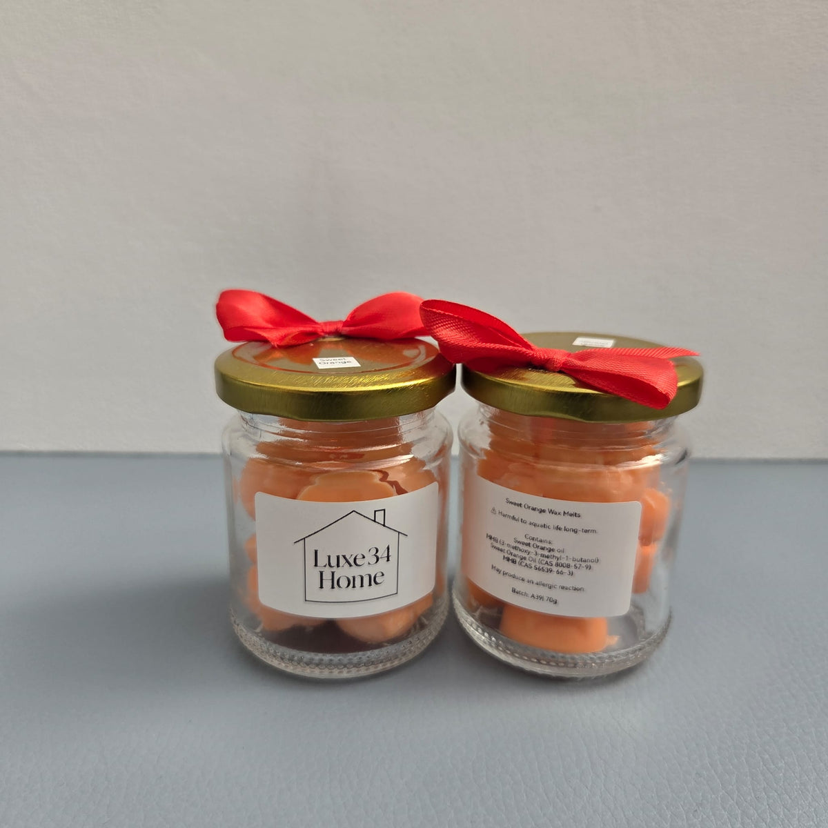 Two Luxe 34 Home sweet orange wax melt jars showing front and back labels with gold lids and red ribbons