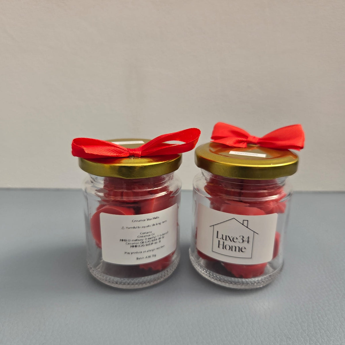 Two Luxe 34 Home cinnamon wax melt jars showing front and back labels with deep red wax melts, gold lids and red ribbons