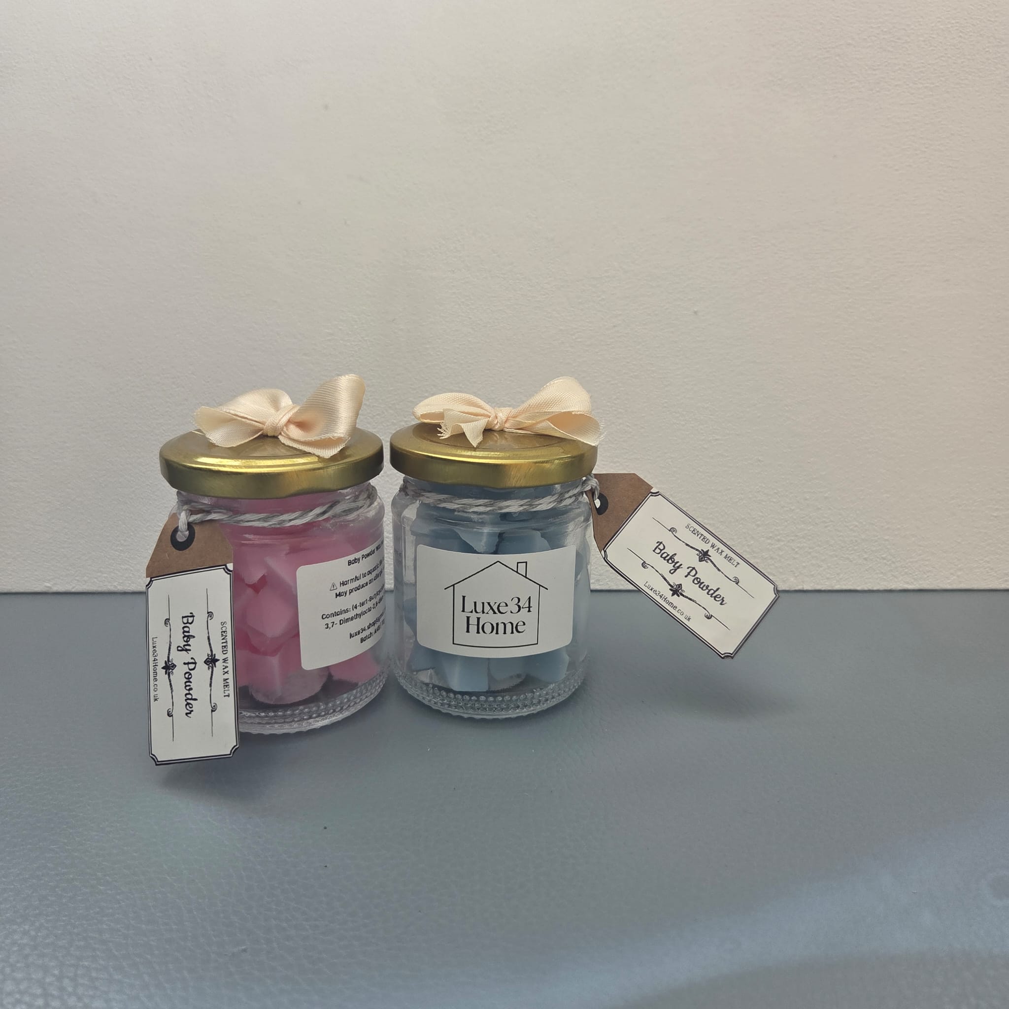 Two Baby Powder wax melt jars side by side — pink and blue — with gold lids, cream ribbon bows and Luxe34 Home labels